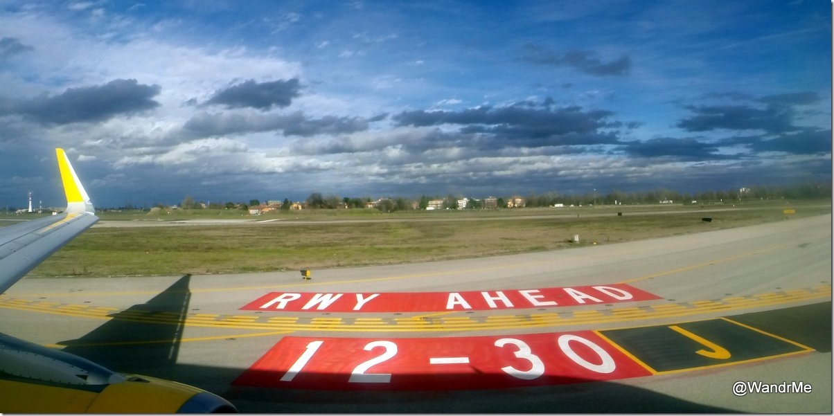 The image shows a view from an airplane window, looking out over an airport runway. The wing of the airplane is visible on the left side of the image. On the ground, there is a large red and white sign that reads "RWY AHEAD" and "12-30 J." The background features a grassy area, some buildings, and a partly cloudy sky.
