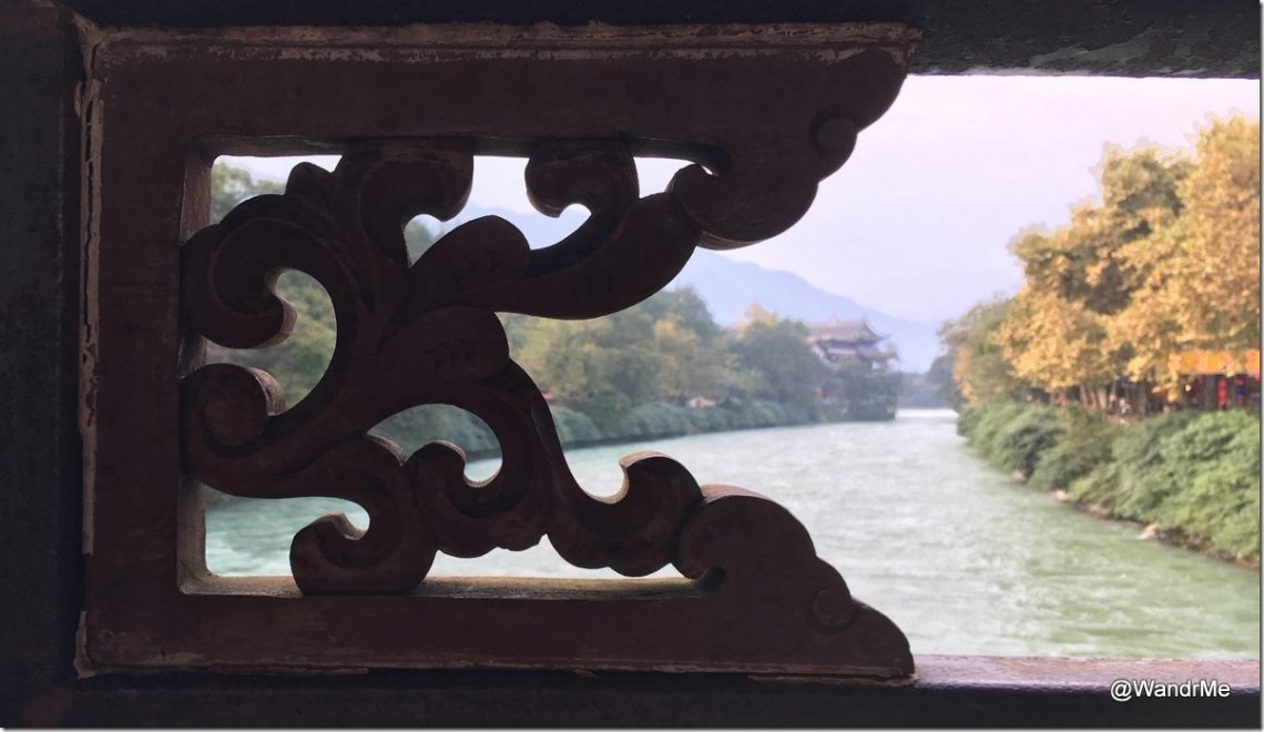 The image shows a scenic view of a river framed by an intricately carved wooden window. The river is flanked by lush green trees on both sides, with some trees displaying autumn colors. In the background, there are mountains and a traditional building with a pagoda-style roof. The sky is slightly overcast, adding a soft light to the scene. The photo is taken from inside a structure, looking out through the decorative wooden frame.
