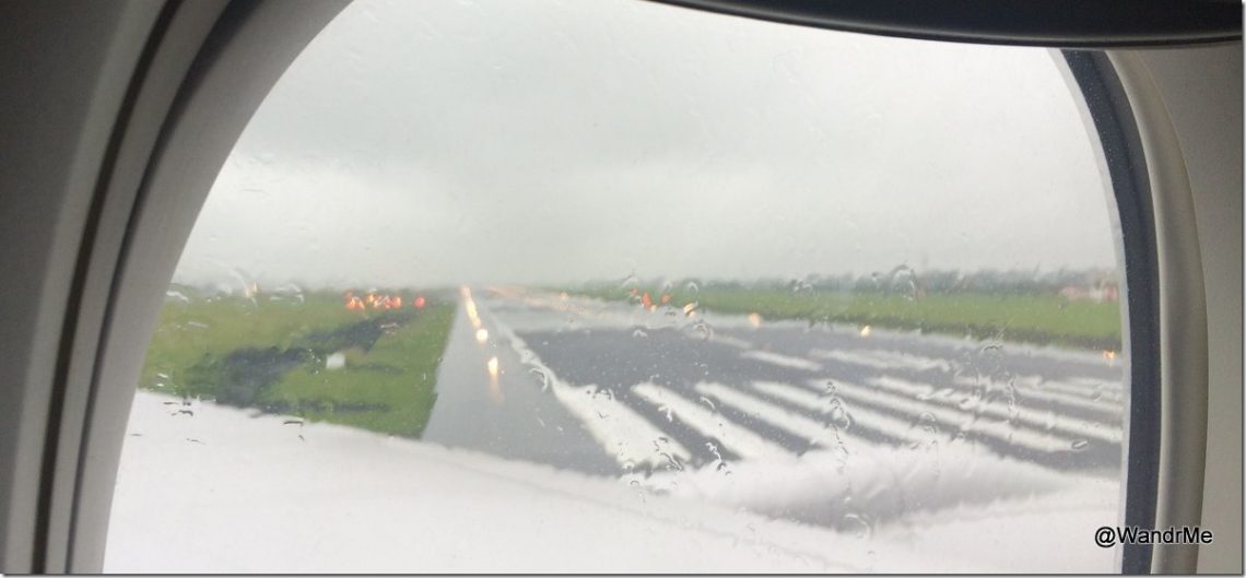 The image shows a view from an airplane window during a rainy day. The airplane is on a runway, which is wet and has visible markings. Raindrops are seen on the window, and the sky is overcast with gray clouds. The runway extends into the distance, and there is green grass on either side of it.