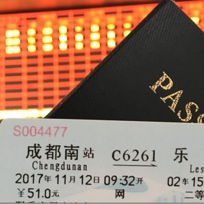 A close-up image of a train ticket and a passport. The train ticket is for a journey from Chengdu South (Chengdunan) to Leshan, scheduled for November 12, 2017, at 09:32 AM. The ticket price is ¥51.0, and the seat number is 02 Car 15B. The background shows a blurred electronic departure board with red and yellow lights.