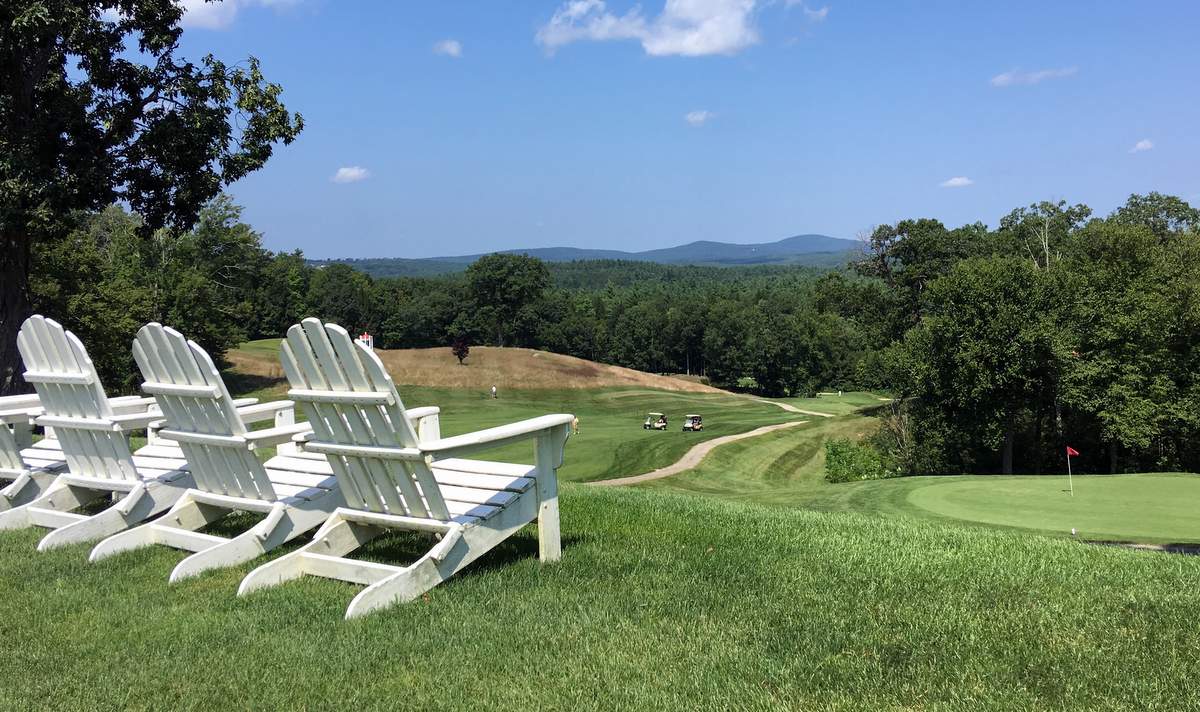 The image shows a scenic view of a golf course with rolling green hills and a clear blue sky. In the foreground, there are four white Adirondack chairs facing the golf course. The course features a well-maintained green with a red flag, a winding path, and several golf carts in the distance. The background includes lush trees and distant mountains.
