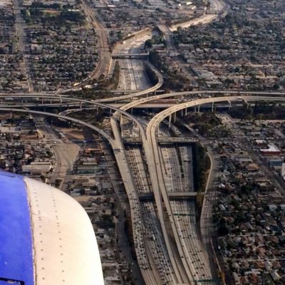 The image shows an aerial view of a complex highway interchange in an urban area. Multiple layers of roads and overpasses intersect, creating a network of ramps and lanes. Surrounding the interchange are densely packed residential and commercial buildings. The wing of an airplane is visible in the lower left corner of the image, indicating that the photo was taken from an aircraft.