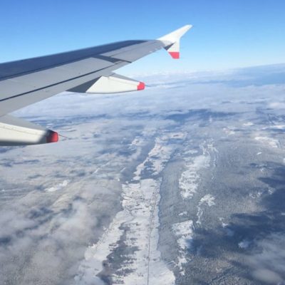 Alt text: Aerial view from an airplane window showing the wing and engine of the aircraft. Below, a vast landscape of snow-covered terrain and forests is visible, with patches of clouds scattered across the scene. The sky above is clear and blue.