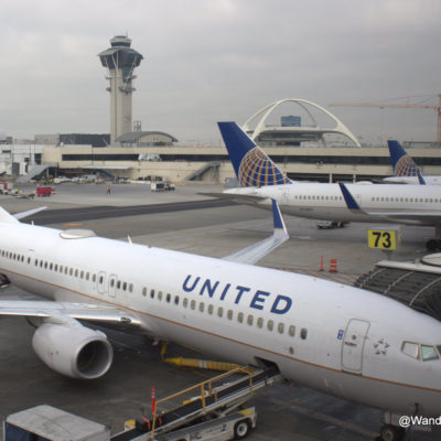 The image shows several United Airlines airplanes parked at an airport gate. The foreground features a United Airlines aircraft with the word "UNITED" clearly visible on its fuselage. In the background, there are more United Airlines planes, an airport control tower, and the distinctive architecture of the Theme Building at Los Angeles International Airport (LAX). The sky is overcast, and various airport ground service vehicles and personnel are visible on the tarmac.