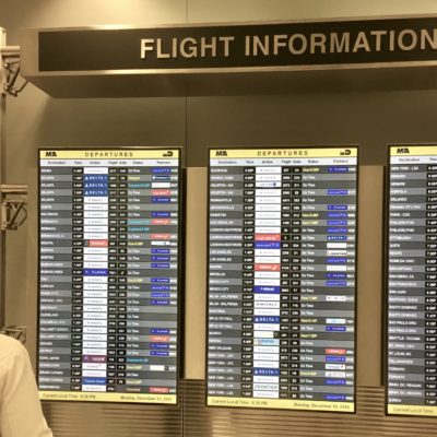 The image shows a person looking at a large flight information display board at an airport. The board is divided into three sections, each listing flight departures. Each section includes columns for destination, time, airline, flight number, gate, status, and partners. The top of the display reads "FLIGHT INFORMATION." The current local time and date are shown at the bottom of the display as 6:26 PM on Monday, December 10, 2018.