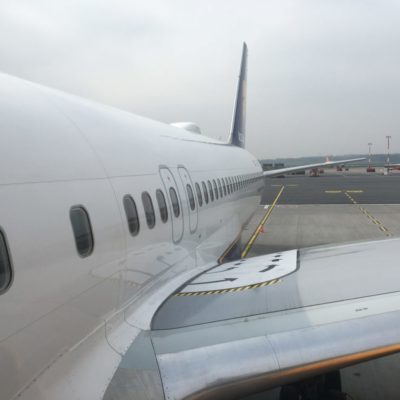 Alt text: A view of an airplane from the side, showing the row of windows and part of the wing. The aircraft is parked on the tarmac at an airport, with the runway and some airport vehicles visible in the background. The sky is overcast.