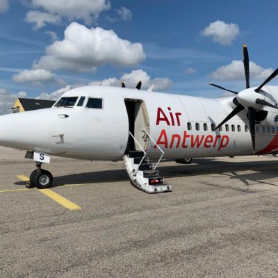 The image shows a white propeller airplane on the tarmac with the words "Air Antwerp" written on the side in red letters. The aircraft has a set of stairs leading up to an open door near the front. The sky is partly cloudy, and there are other buildings and aircraft visible in the background.