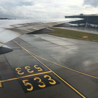 The image shows the view from an airplane window, focusing on the wing of the aircraft and the tarmac below. The tarmac has yellow markings, including an arrow and the numbers "533" and "333." In the background, there is an airport terminal building with glass windows and a jet bridge connected to another airplane. The sky is cloudy, suggesting overcast weather.