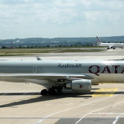 A Qatar Airways airplane is being towed on the tarmac at an airport. The aircraft is white with the Qatar Airways logo and text in both English and Arabic. In the background, another airplane, belonging to Air France, is taxiing on the runway. The sky is partly cloudy, and there are some buildings and greenery visible in the distance.