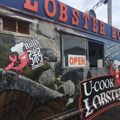 The image shows a food truck with a sign on top that reads "LOBSTER ROLLS" in large red letters. The side of the truck features a cartoon lobster wearing a chef's hat, with text that says "Rolls! Congee" and "Become a LOBSTER CHEF in 12 Minutes!" There is also a sign that says "U-COOK LOBSTER." The truck has a window with an "OPEN" sign displayed. The background of the truck is designed to look like a rocky surface.