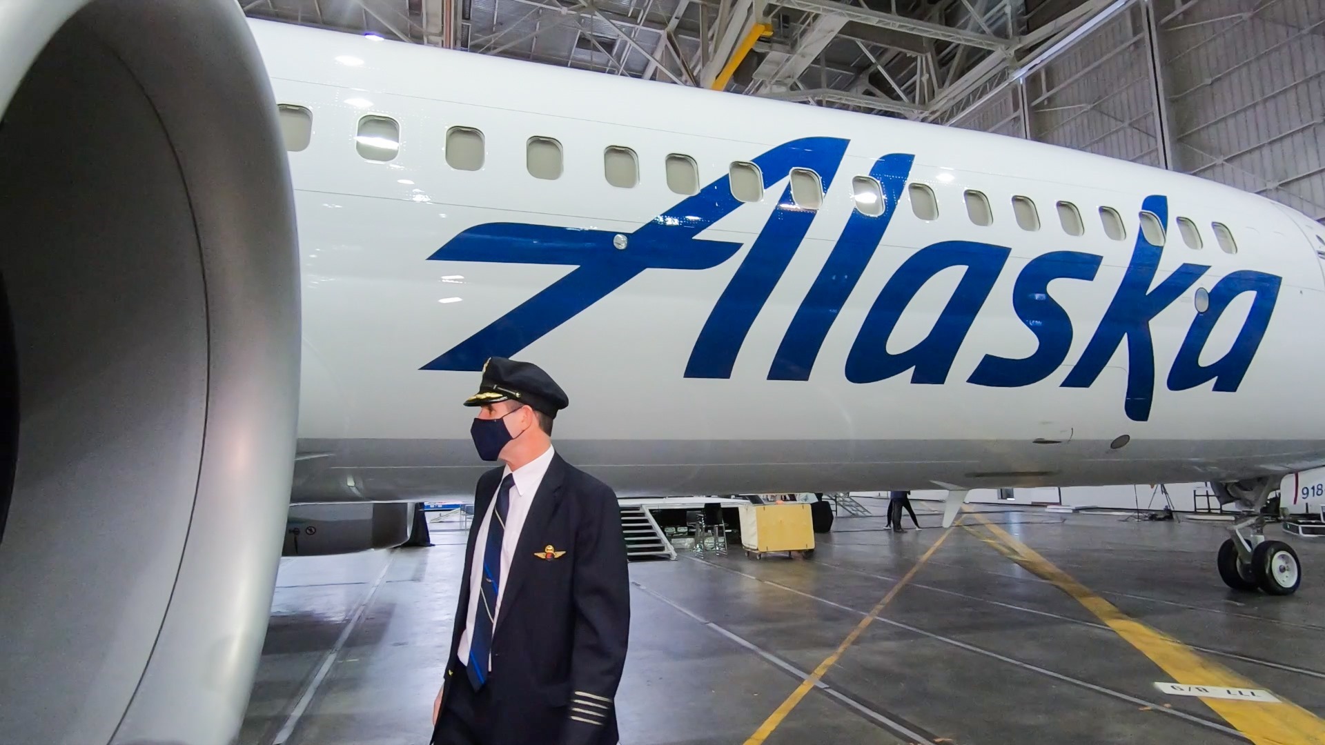A pilot wearing a uniform and a face mask is standing in front of an Alaska Airlines airplane inside a hangar. The airplane has the word "Alaska" prominently displayed on its side. The hangar is spacious with various equipment and machinery visible in the background.