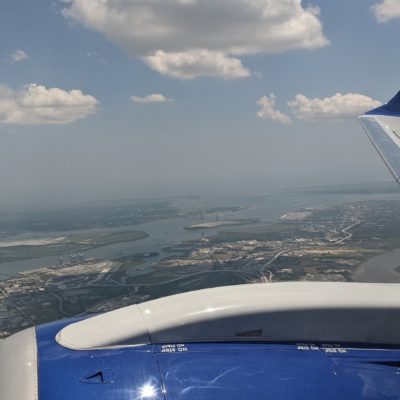 Aerial view from an airplane window showing the aircraft's wing and engine. Below, there is a landscape with a river, bridges, and an urban area with buildings and roads. The sky is partly cloudy with scattered white clouds.