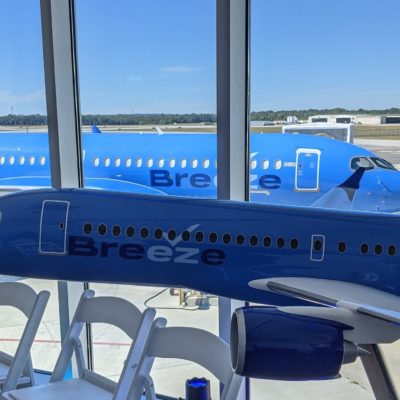 The image shows a large blue airplane parked on the tarmac outside an airport terminal. In the foreground, there is a model of a blue airplane with the word "Breeze" written on its side, placed on a stand near some white chairs. The model airplane is positioned in front of large windows that provide a clear view of the actual airplane outside. The sky is clear and blue, and the airport runway and surrounding area are visible in the background.