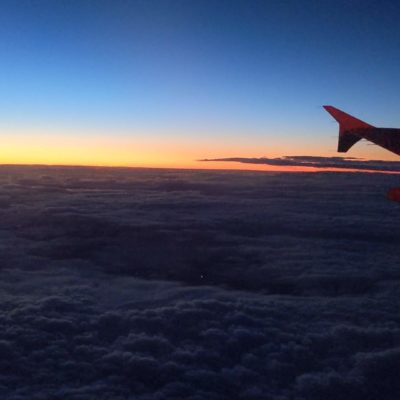 The image shows a view from an airplane window during sunset or sunrise. The sky transitions from a deep blue at the top to a warm orange near the horizon. Below, a blanket of clouds stretches out, with the airplane wing visible on the right side of the image.