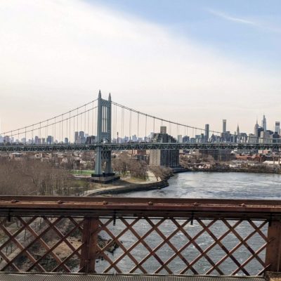 Alt text: A suspension bridge spans across a river with a city skyline in the background. The bridge has tall towers and cables, and the cityscape features numerous high-rise buildings. In the foreground, there is a metal fence with a crisscross pattern. The sky is mostly clear with a few clouds.