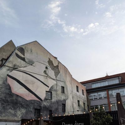 A large mural is painted on the side of a building, depicting a person in a hat holding a large kite or sail. The mural features muted colors and abstract elements, including a map-like shape and several small human figures. The building is part of a street scene with other buildings, one of which has a distinctive tower with a weather vane. String lights are strung across the foreground, suggesting an outdoor seating area or café. The sky is partly cloudy.