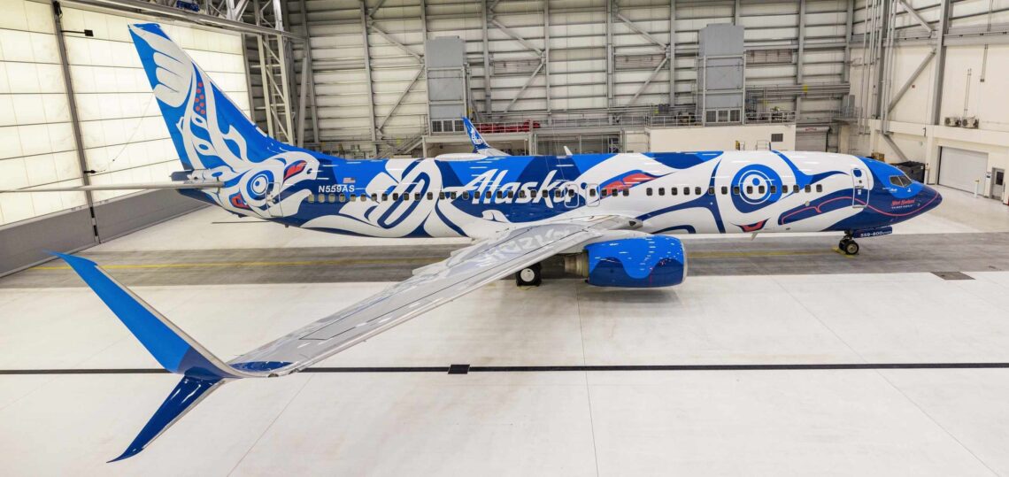 The image shows an Alaska Airlines airplane inside a hangar. The aircraft is painted with a special livery featuring bold blue and white designs inspired by Northwest Coast Native art. The word "Alaska" is prominently displayed on the side of the plane. The tail and winglets also feature intricate designs in blue and white. The hangar is spacious and well-lit, with the airplane positioned centrally.