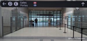 The image shows an airport terminal with a sign overhead indicating directions to gates D and E. The sign includes a Canadian flag and a globe symbol. A person is standing in the middle of the hallway with a suitcase, looking at their phone. The area is enclosed with glass walls, and there are signs for restrooms in the background. The floor is tiled, and there are barriers guiding the path.