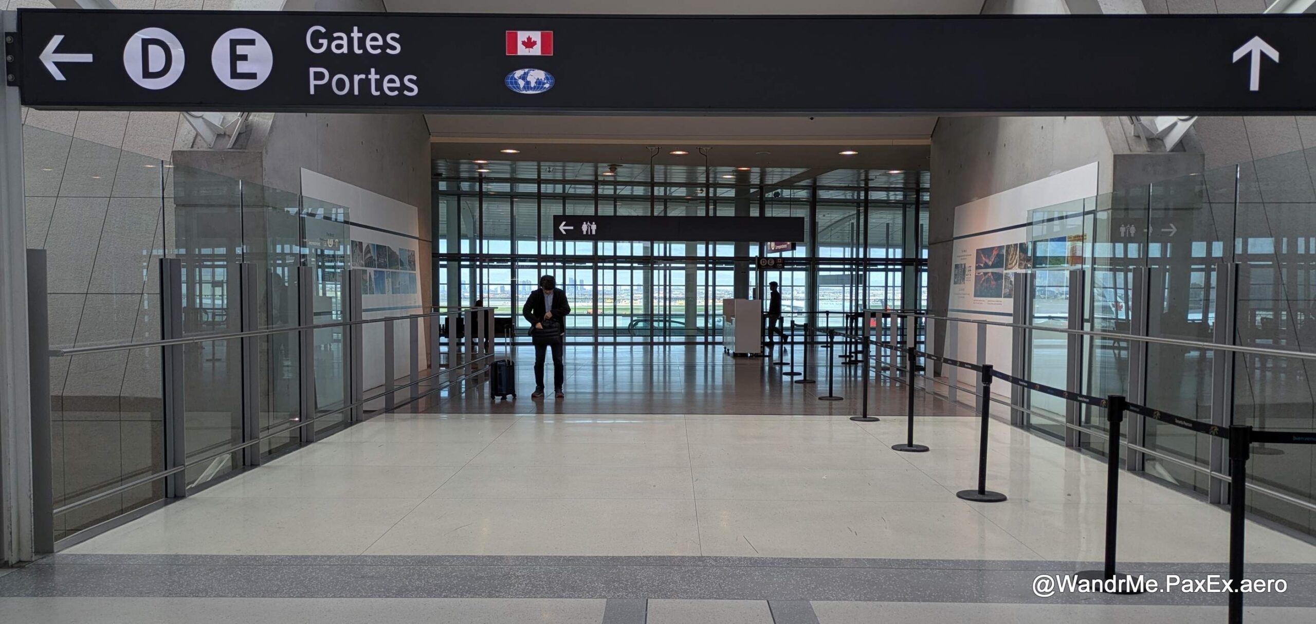The image shows an airport terminal with a sign overhead indicating directions to gates D and E. The sign includes a Canadian flag and a globe symbol. A person is standing in the middle of the hallway with a suitcase, looking at their phone. The area is enclosed with glass walls, and there are signs for restrooms in the background. The floor is tiled, and there are barriers guiding the path.
