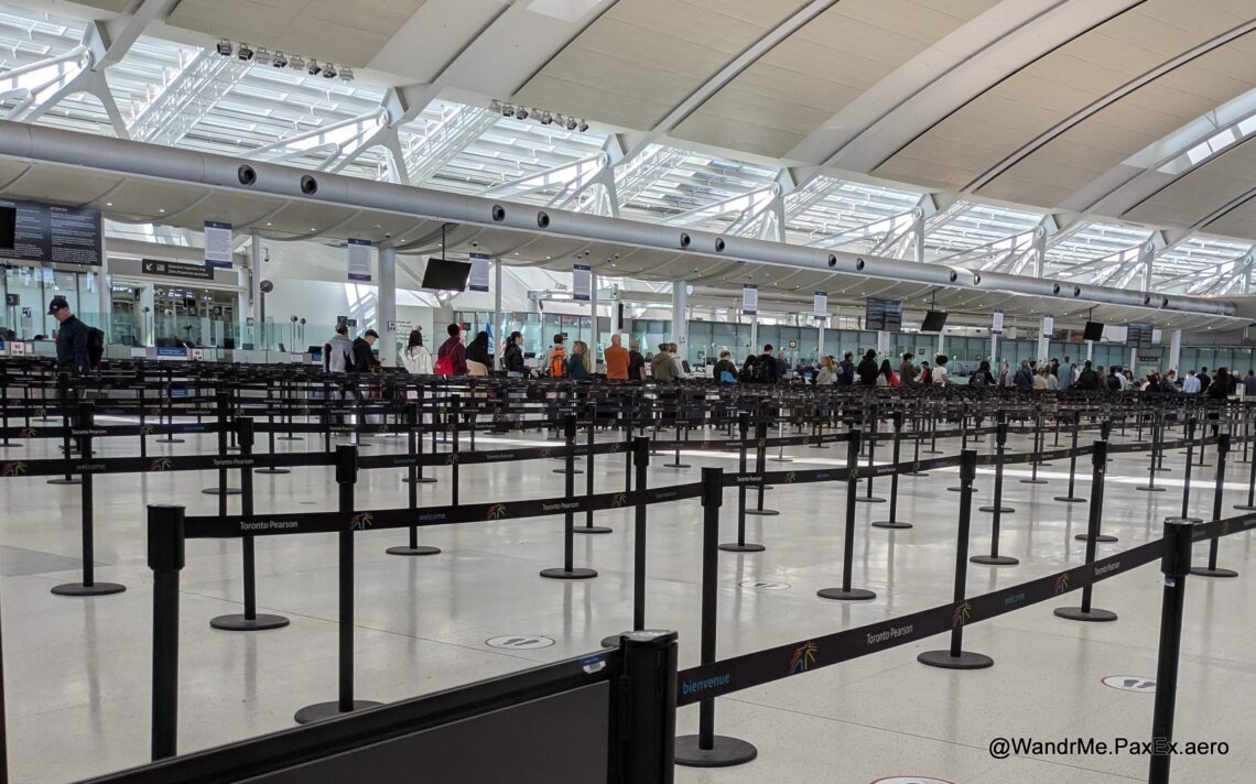 The image shows a large, open area inside an airport terminal with multiple rows of stanchions and retractable belts forming a queue system. There are people standing in line, and the ceiling features a modern design with large windows allowing natural light to enter. Signs indicate that this is Toronto Pearson Airport.