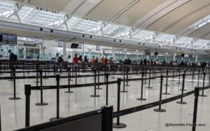 The image shows a large, open area inside an airport terminal with multiple rows of stanchions and retractable belts forming a queue system. There are people standing in line, and the ceiling features a modern design with large windows allowing natural light to enter. Signs indicate that this is Toronto Pearson Airport.