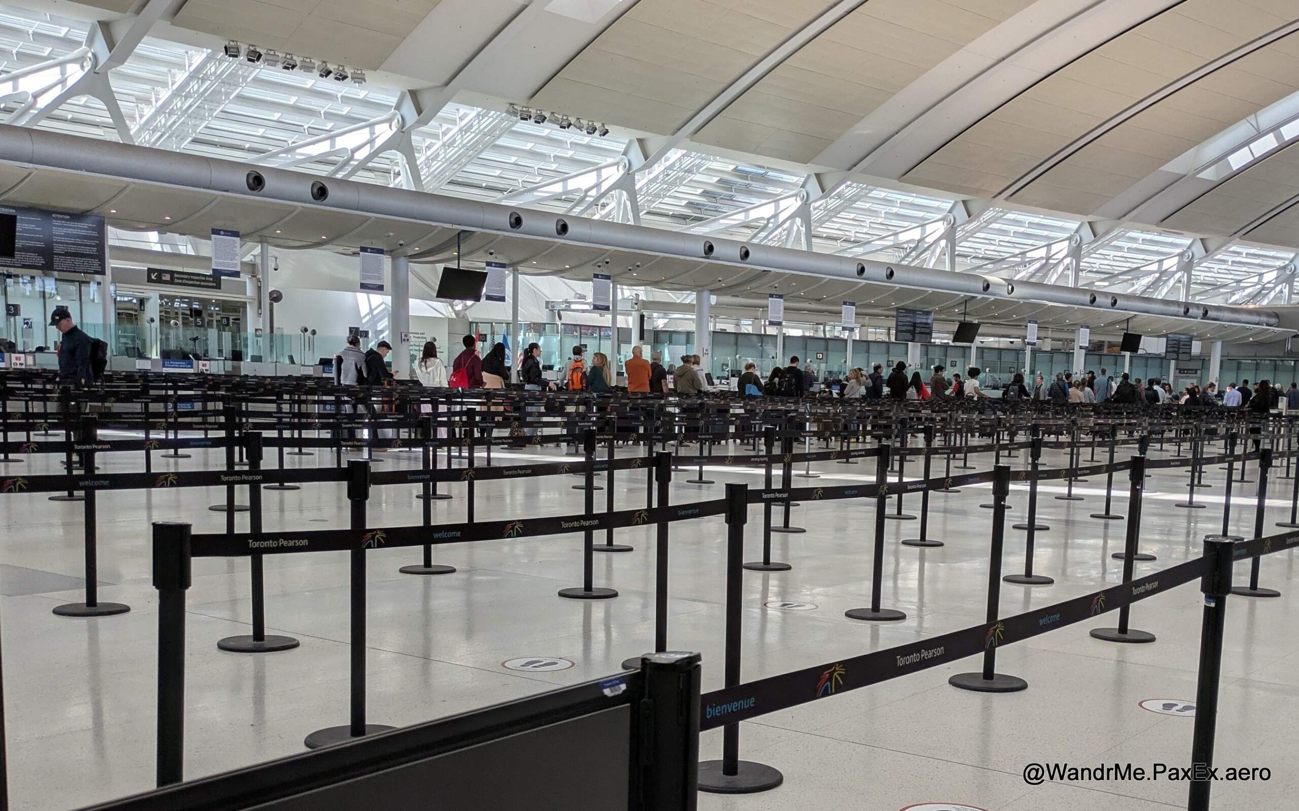 The image shows a large, open area inside an airport terminal with multiple rows of stanchions and retractable belts forming a queue system. There are people standing in line, and the ceiling features a modern design with large windows allowing natural light to enter. Signs indicate that this is Toronto Pearson Airport.
