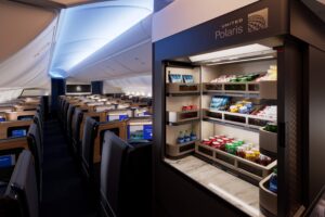 The image shows the interior of an airplane cabin, specifically the business class section. On the right, there is a well-stocked snack and beverage station labeled "United Polaris," featuring various drinks and snacks. The cabin has spacious seating with individual screens, and the lighting is soft and ambient, creating a comfortable atmosphere.