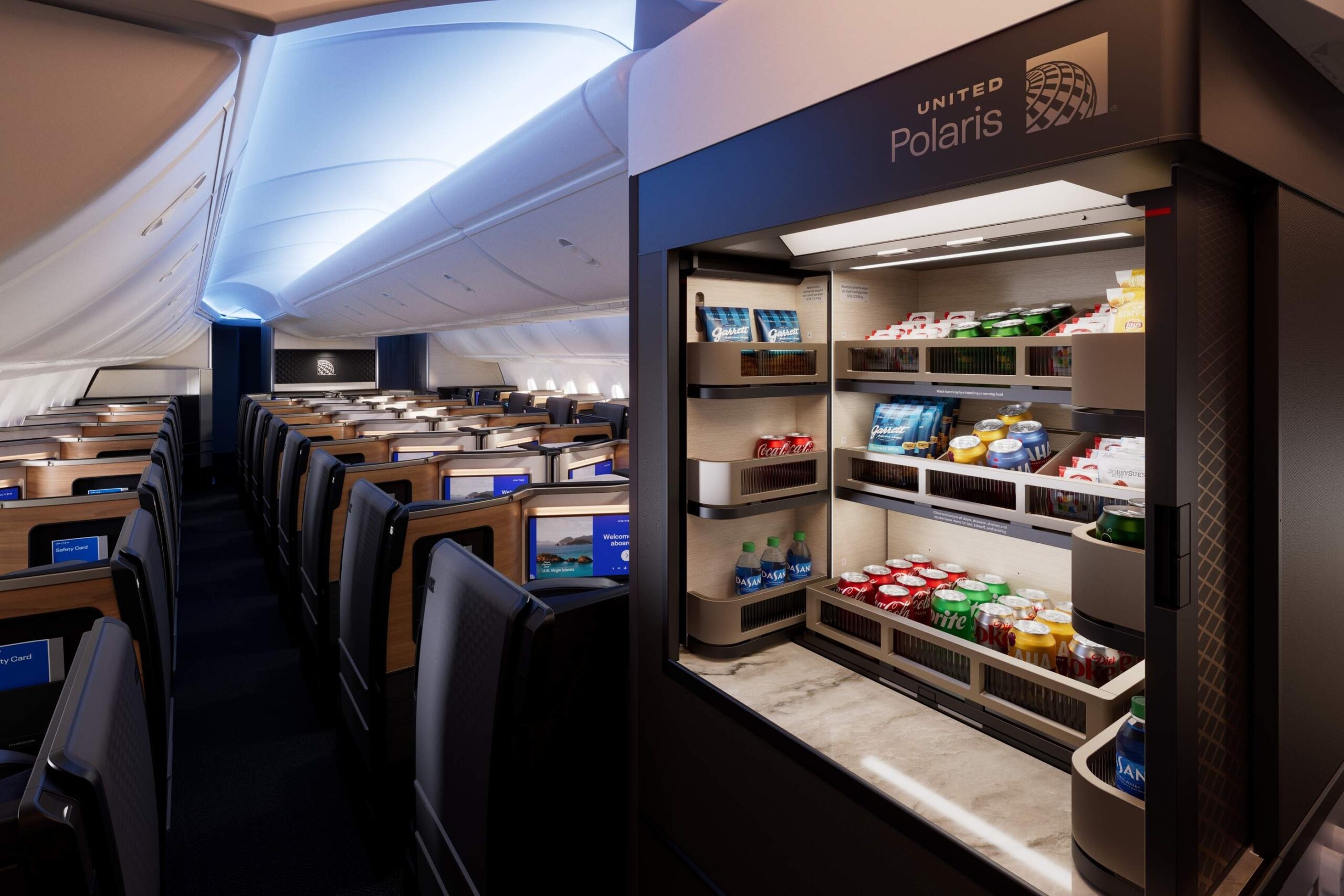 The image shows the interior of an airplane cabin, specifically the business class section. On the right, there is a well-stocked snack and beverage station labeled "United Polaris," featuring various drinks and snacks. The cabin has spacious seating with individual screens, and the lighting is soft and ambient, creating a comfortable atmosphere.