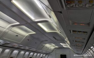 The image shows the interior of an airplane cabin, focusing on the overhead compartments and ceiling. The compartments are closed, and the ceiling has illuminated panels. There are also air vents and light controls visible. The cabin appears to be empty or sparsely occupied, with a few heads visible at the bottom of the image.