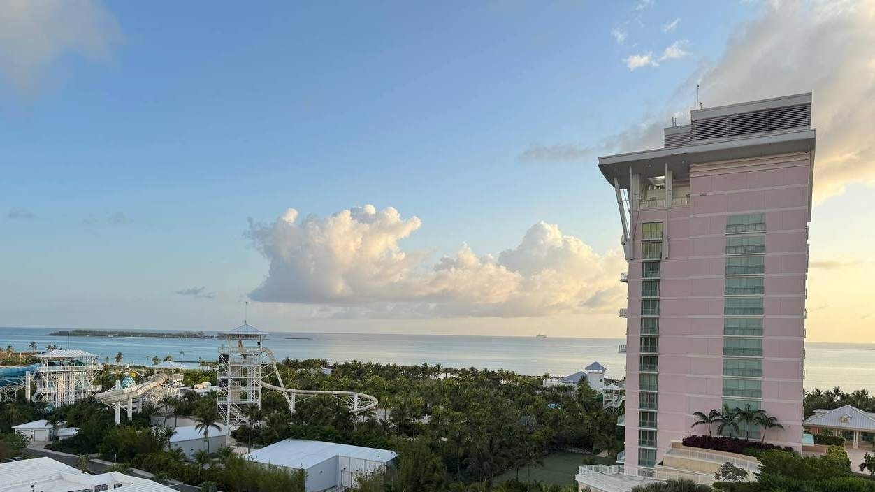 A tall pink building stands on the right side of the image, overlooking a scenic view of the ocean. In the foreground, there are lush green trees and a water park with slides and structures. The sky is mostly clear with a few fluffy clouds, and the ocean stretches out to the horizon.