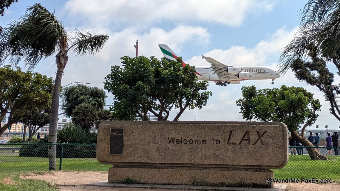 A380 landing at LAX, seen from a park with a sign and some trees.