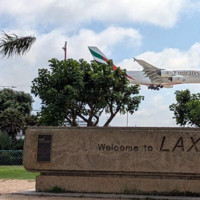A380 landing at LAX, seen from a park with a sign and some trees.